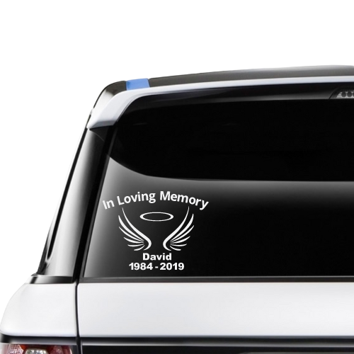 A custom vinyl sticker with the text 'In Loving Memory insert name adhered to the rear window of a vehicle. The sticker includes an illustration of wings and a halo.